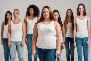 Fototapeta premium Portrait of beautiful young caucasian plus size woman in white shirt looking at camera. Group of diverse women posing, standing isolated over grey background