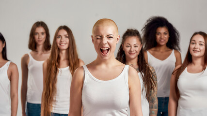 Portrait of cheerful young caucasian woman with shaved head in white shirt winking at camera. Group of diverse women standing isolated over grey background