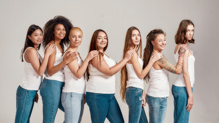 Group of beautiful diverse young women wearing white shirt and denim jeans looking at camera while posing, standing isolated over grey background
