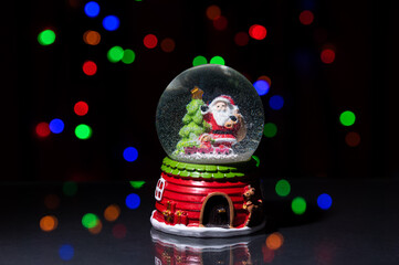 A snow globe with a Christmas tree and Santa Claus on a dark, glossy table against a background of festive lights.