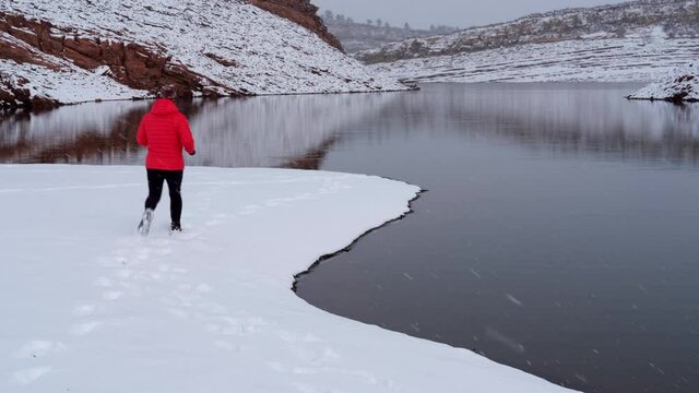 senior male in a down jacket is jogging along a shore of Horsetooth Reservoir at foothills of Rocky Mountains in northern Colorado, winter scenery with falling snow