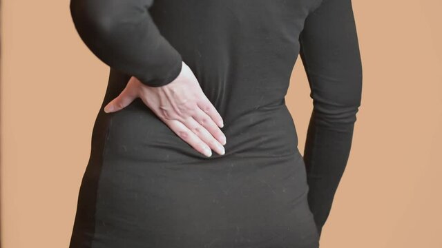 The woman rubs her back. A woman with back pain stands against a light wall.