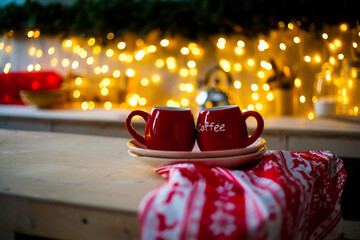 there are a couple of coffee cups and saucers on a wooden table on a towel,and lights are burning in the background