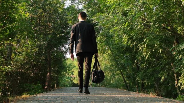 A man in a business suit walks along the road in the park with a travel bag in his hand. Goes with his back to the video camera, shooting 120 fps.