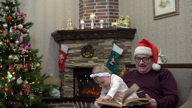 Grandfather in a Santa hat put his granddaughter on his knees and reads a fairy tale by the fireplace before Christmas. Festive atmosphere before the new year