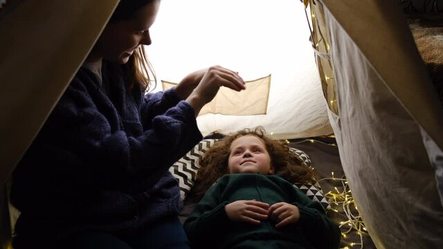 Happy caring mother with daughter playing in puppet theater of shadows, sitting in tent decoration string lights. Social distancing and self isolation in quarantine lockdown for Coronavirus Covid 19
