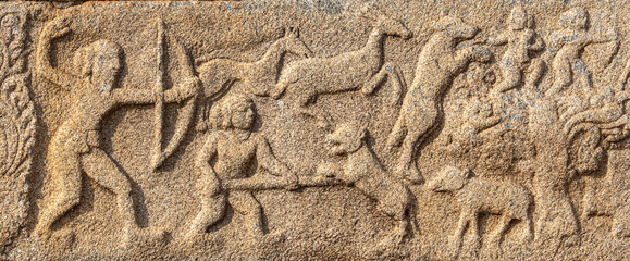 Hampi, Karnataka, India - November 4, 2013: Mural sculpture on brown stone at Royal Enclosure. Closeup of hunting scene with people and deer. Bow and spear as weapons.