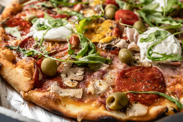 Baked pizza with salami, prosciutto, olives and arugula with red sauce and melted cheese on the table, close up