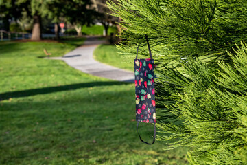 Handmade fabric face mask with a holiday print, hanging on an evergreen tree branch in a park
