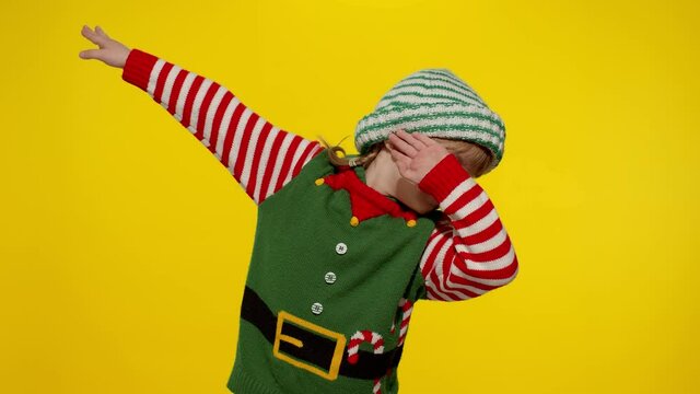 Little fun blonde kid teen teenager girl in Christmas elf Santa helper costume performing dab dance on yellow background. Child dancing making dabbing movement with hands up, internet meme gesture