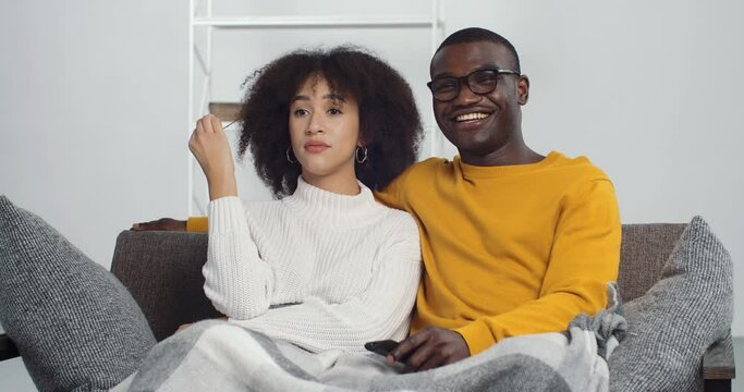 Dissatisfied African American Woman Ethnic Girl Sitting On Couch With Her Smiling Black Man Husband Feeling Boredom, Guy Switching Channels To TV Changing Program Film. Young Family Resting At Home