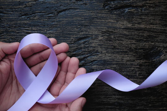 Top view of hands holding lavender or light purple orchid color ribbon on dark wooden background. General and testicular cancer and epilepsy awareness, national cancer prevention month.