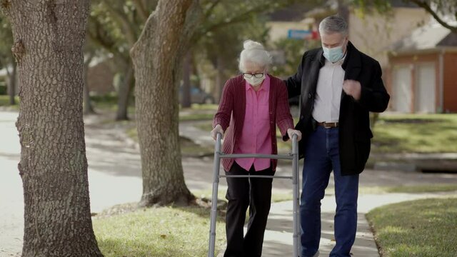 A man helps an elderly relative who needs to use a walker enjoy a walk outdoors both wearing masks as a precaution due to COVID-19.