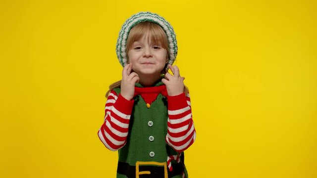Christmas wish concept. Little fun blonde kid teen teenager girl in Christmas elf Santa helper costume hopes for dreams come, making a wish, fingers crossed, prays for better life. Shy smiling child