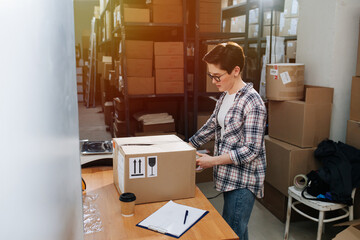 Busy female warehouse worker closing box with cello tape, sealing it. She has short hair, wearing checkered shirt and glasses.