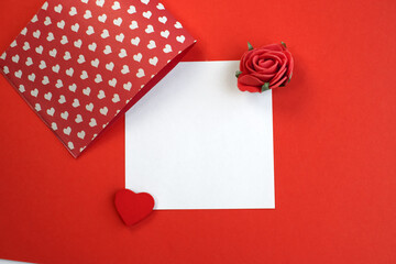 A white square leaf with a heart and a rose lies on a red background. Holiday background template with copy space