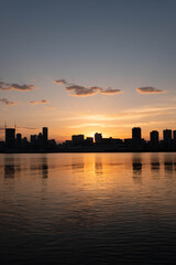 夜明け前、大阪淀川河川敷から梅田の高層ビルをのぞむ。オレンジ色に染まった空に摩天楼がシルエットになり水面に映る