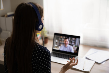 Rear view woman wearing headphones using laptop, making video call, businesswoman working at home,...