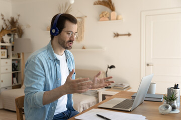 Confident man wearing glasses and headphones making video call, using laptop, sitting at work desk, home office, engaged in online negotiations, internet meeting, mentor coach leading webinar