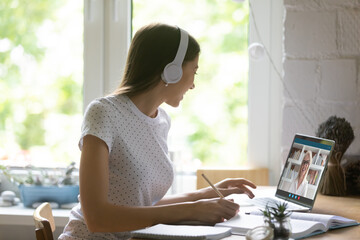 Close up young woman wearing headphones studying online with mature teacher, using laptop, female student making video call to mentor coach, listening to lecture, writing notes, watching webinar