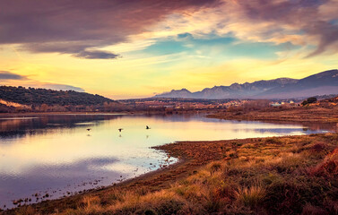 Lake landscape at sunset with ducks flying flush with the water and dramatic sky.
