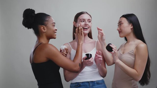 Funny multi-cultural girls young women using cosmetic ream applying product on their faces having fun together in beauty studio advertising background.