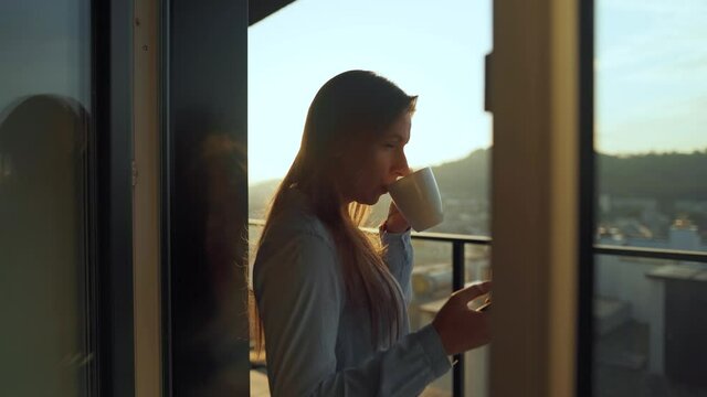 Woman starts her day with a cup of tea or coffee and checking emails in her smartphone on the balcony at dawn.