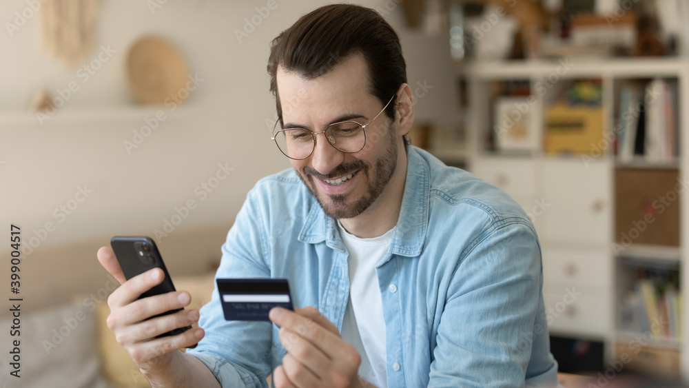 Wall mural close up smiling man wearing glasses making secure internet payment, holding smartphone and plastic 