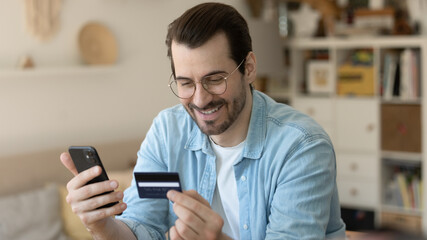 Obraz na płótnie Canvas Close up smiling man wearing glasses making secure internet payment, holding smartphone and plastic credit or debit card, customer purchasing, shopping online at home, client browsing banking service