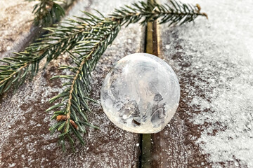 Frozen bubble with ice crystals and spruce branch on icy wooden bench in park.Christmas background