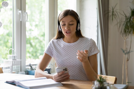 Woman wearing headphones speaking, holding smartphone, chatting with friends or colleagues, sitting at desk at home, student learning language, engaged in conference, using social media apps