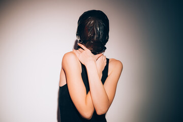 Female abuse. Domestic violence. Social isolation. Gender discrimination. Art portrait of assaulted woman with head wrapped in black polyethylene garbage bag strangling throat in spotlight.