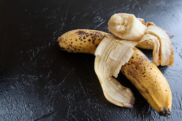 Overripe bananas with dark spots on the skin on black background.   One peeled banana and one...