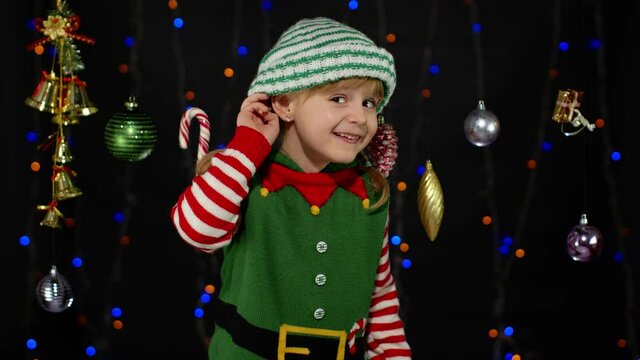 Kid girl in Christmas elf Santa Claus helper costume keeps hand near ear tries to overhear what present gifts parents going to give her. Child interested in someones conversation. New Year holidays