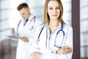 Fototapeta premium Professional beautiful woman-doctor with a stethoscope is standing with crossed arms in a clinic. Young doctors at work in a hospital. Medicine and healthcare concept