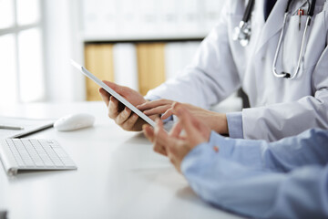 Unknown male doctor and patient woman discussing current health examination while sitting in clinic and using tablet computer, closeup of hands. Medicine and healthcare concept