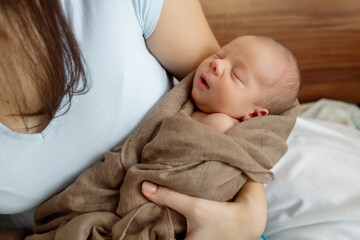 Young mother of a family brunette holds a newborn toddler in her arms - her tiny son in the bedroom at home
