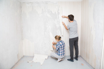 Young adult man and woman tearing off light wallpaper from wall in room. Working together. Making interior change. Back view.