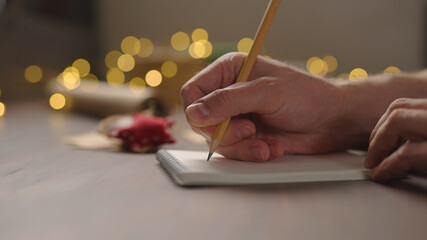 man writes christmas gift list for holidays in notepad with a pencil under warm light in the evening
