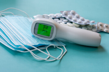 Infrared thermometer with digital green colored screen showing high body temperature on pile of medical masks and pills in blisters. on blue background. Concept of stopping virus illness and influenza