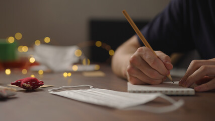 closeup man writes Shopping list in notepad with a pencil under warm light in the evening with medicine mask near hand