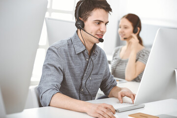 Casual dressed young man using headset and computer while talking with customers online. Group of operators at work. Call center