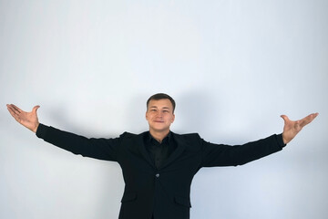 Man is standing on grey background. Showman magician presents his show, spreading his hands up to the sides. He is wearing in black suit. Concept of performances, entertainments, shows and circus.