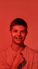Fototapeta premium Portrait of happy teenaged disabled boy with Down syndrome smiling at camera, pointing at himself while posing isolated over red light background