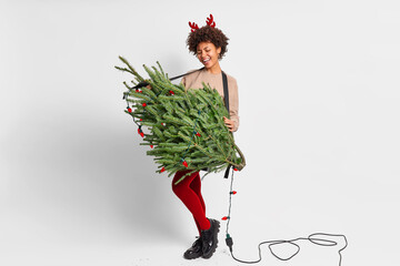 Joyful dark skinned woman has fun and pretends playing guitar on fir tree expresses happiness prepares for New Year concert wears red reindeer antlers stands in full size against white background