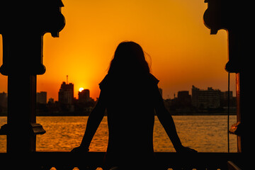 A young woman looking at the orange sunset on the Nile river from a boat, the sun is hiding in the...