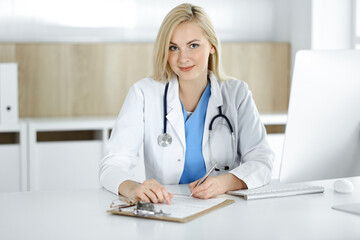 Woman-doctor at work while sitting at the desk in hospital or clinic. Blonde cheerful physician filling up medication history record form