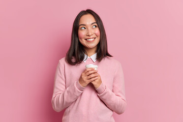 Portrait of brunette Asian woman with dreamy expression smiles pleasantly daydreams with takeaway coffee wears neat pink jumper poses indoor. Pensive female model feels good isolated over rosy wall