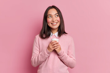 Portrait of brunette Asian woman with dreamy expression smiles pleasantly daydreams with takeaway...