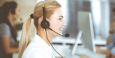 Blonde business woman using headset for communication and consulting people at customer service office. Call center. Group of operators at work